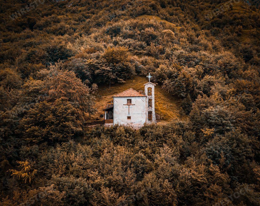 A bird's-eye view of a church standing on top of fall colored hill ,surrounded by a forest at sunset in Dolomites.