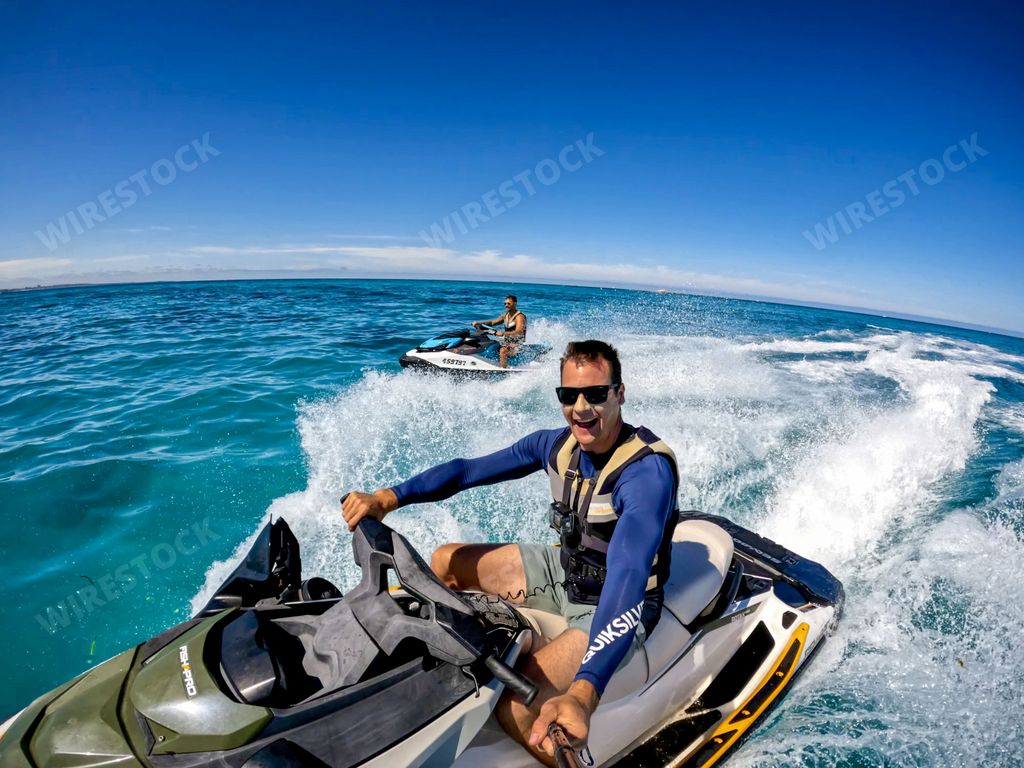 Two men enjoying a sunny day riding jet skis on clear blue ocean water with a bright blue sky in the background.