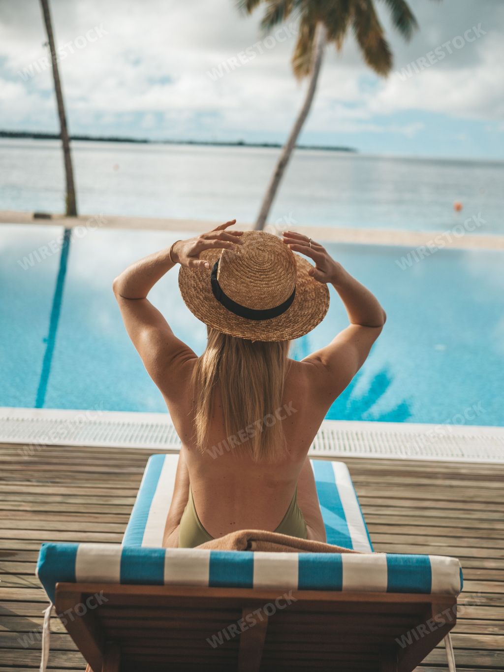 A woman in a straw hat relaxing on a lounge chair by a pool overlooking the ocean on a sunny day.