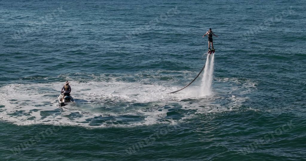 A person enjoying a water jetpack ride while another person assists on a jet ski in the ocean.