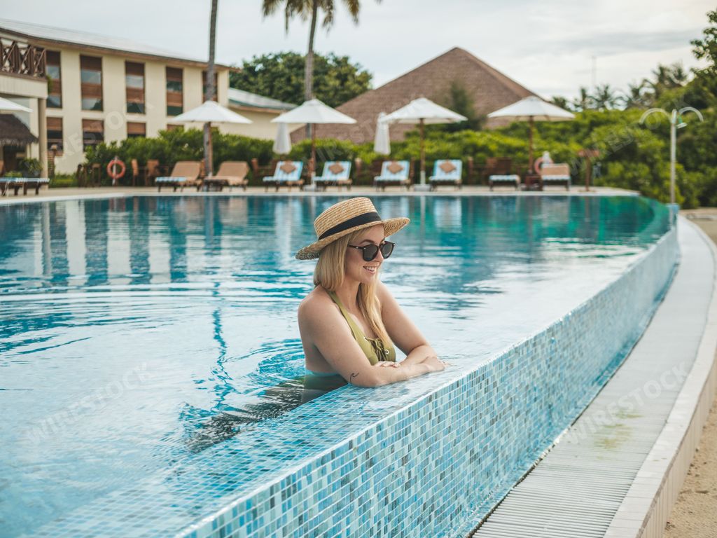 A young woman in a straw hat and sunglasses relaxing in an infinity pool at a tropical resort.