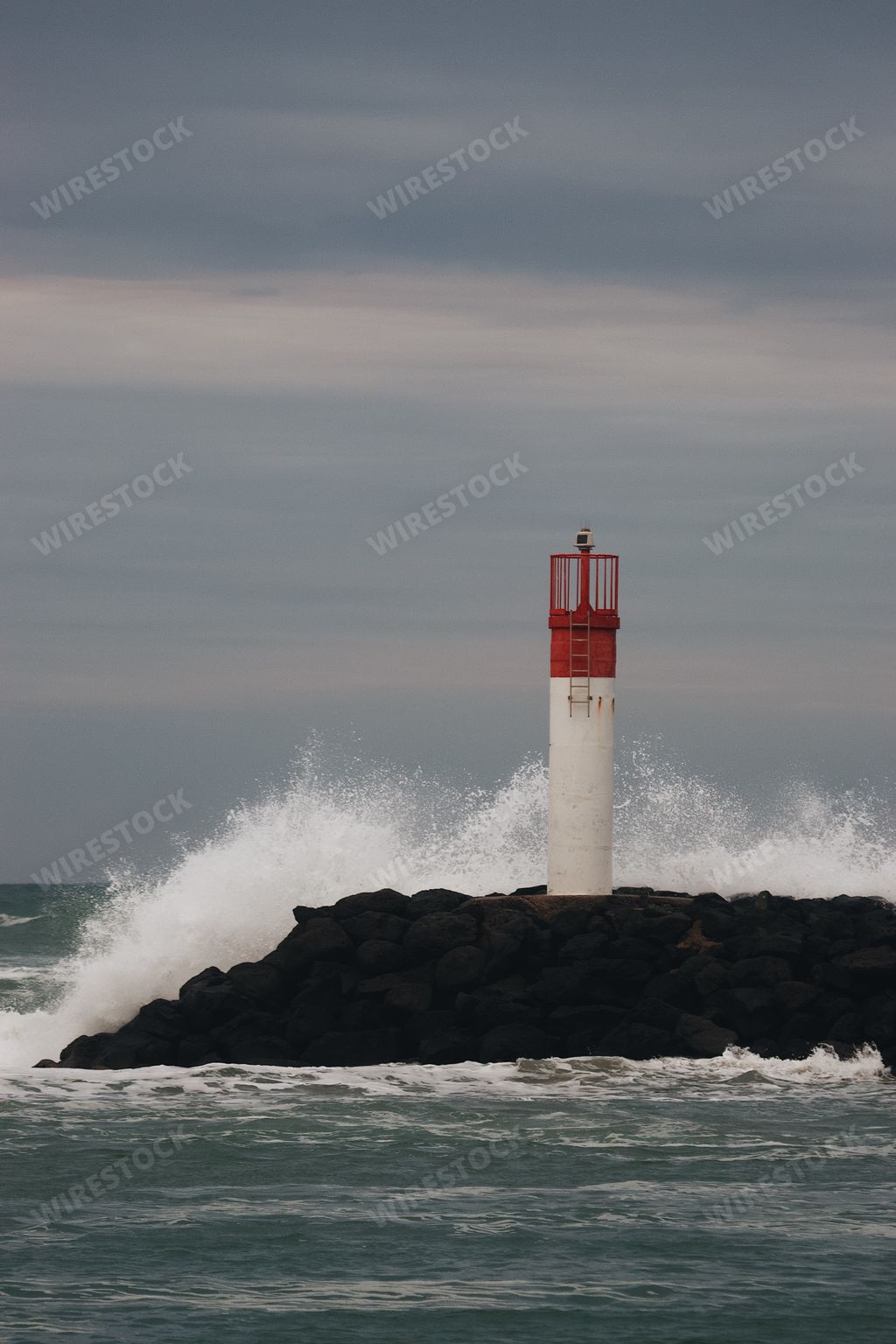 A vertical shot a white lighthouse  with a red top and a wave hitting it