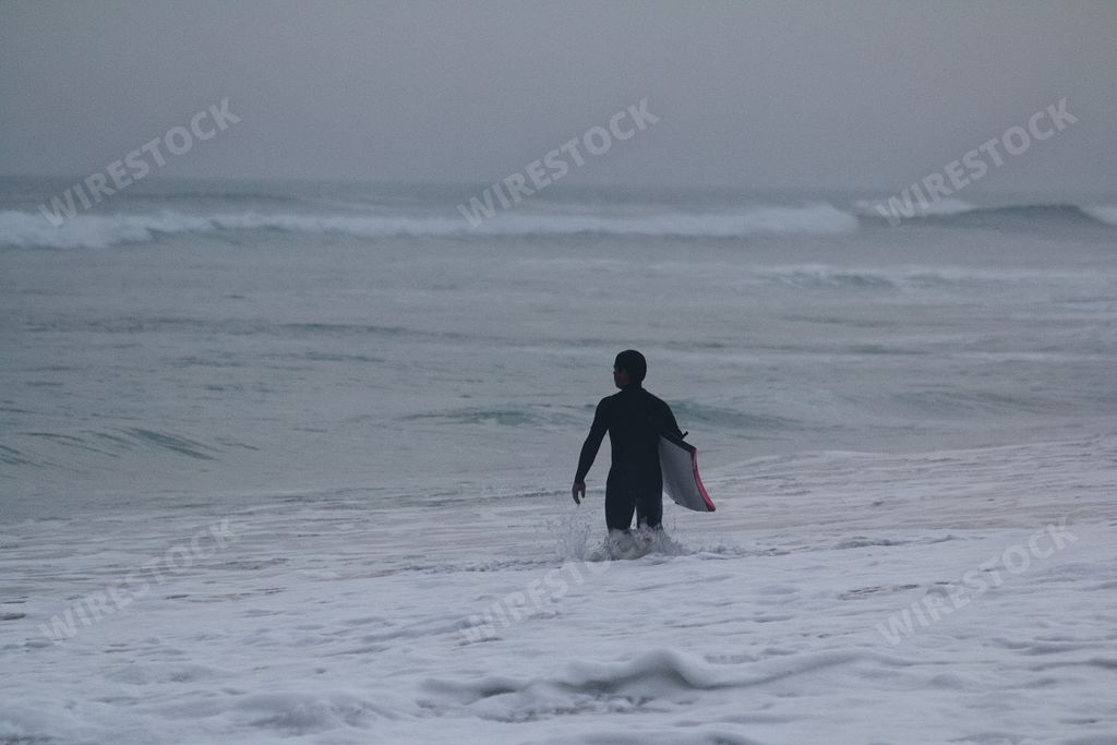 A male surfer entering the water with his surfboard on a gloomy day