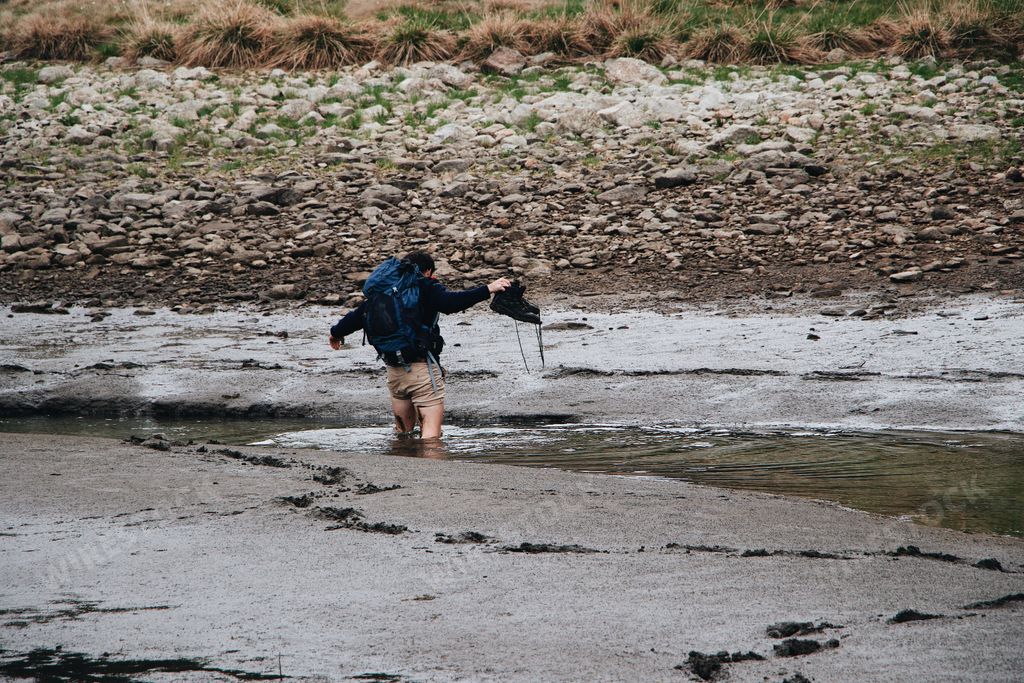 An unhappy traveler with a backpack holding his boots in his hand while soaking in a muddy lake