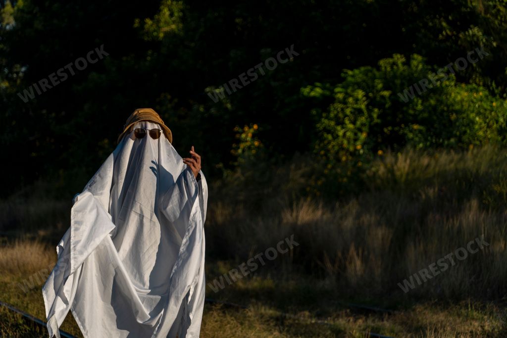 A person in a funny ghost costume with a white sheet, sunglasses, and a yellow hat in a park