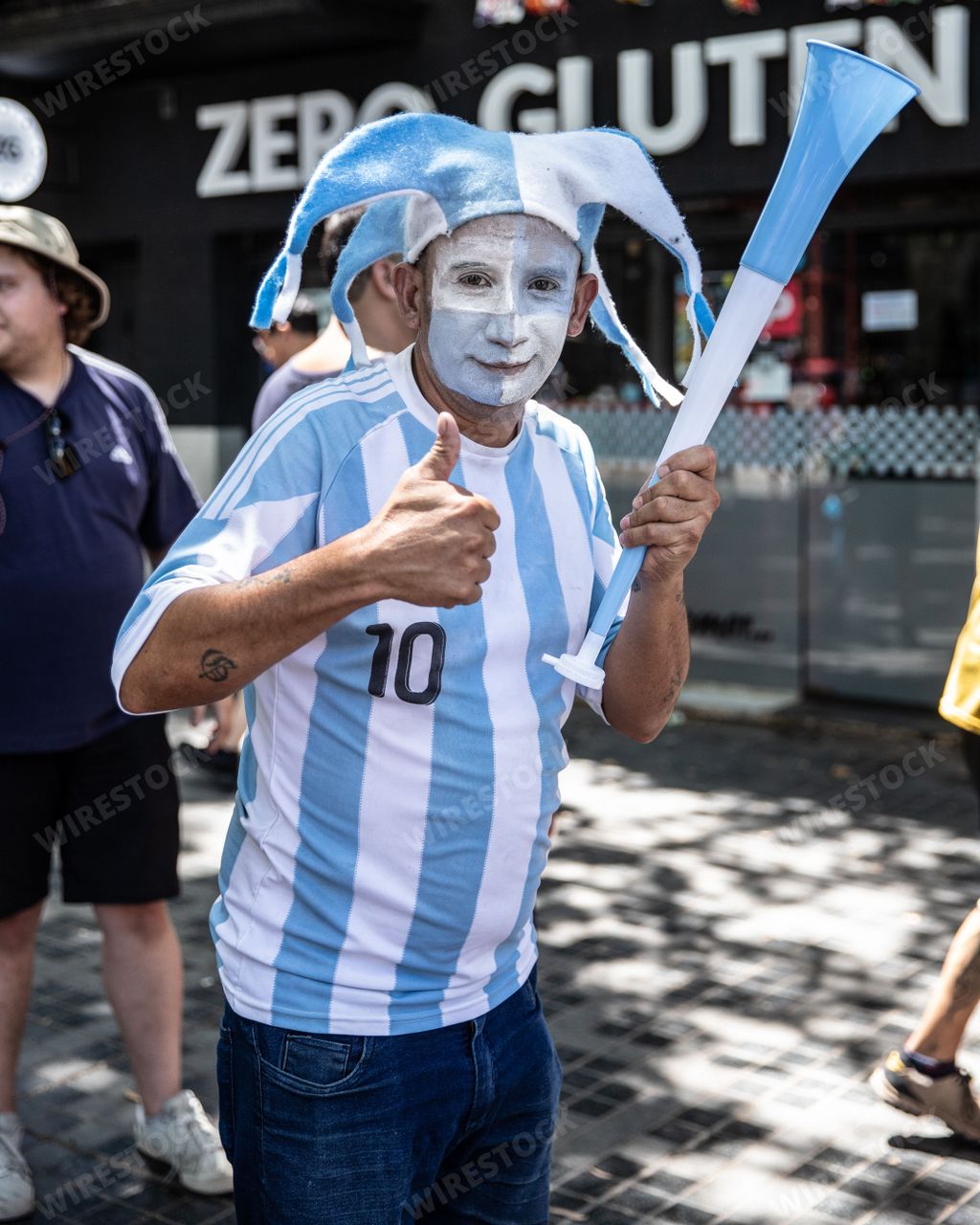 A Caucasian male wearing theatrical face paint holds two blue and white baseball bats in a standing position