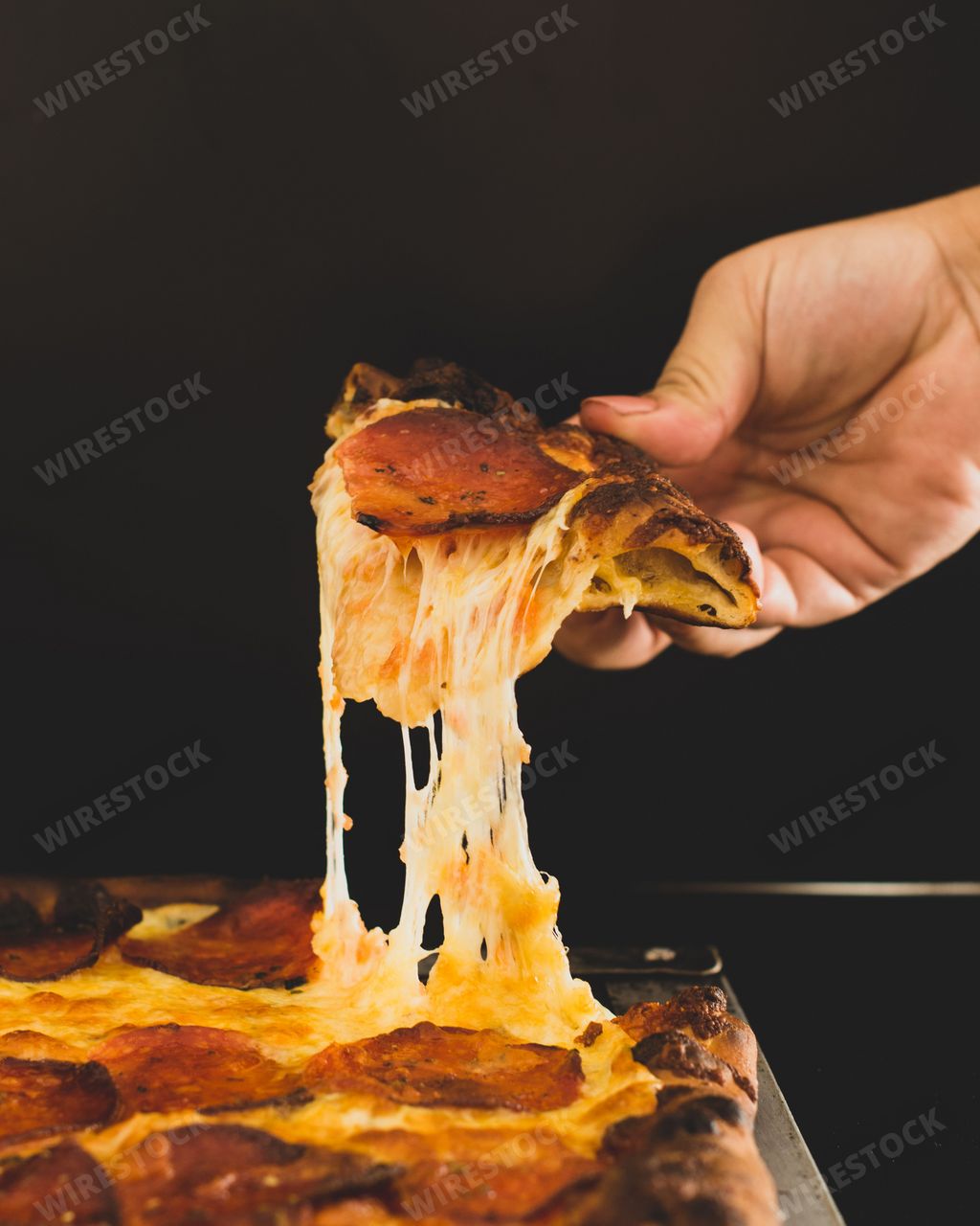 A hand holding a piece of freshly-baked pepperoni pizza, with a slice being lifted up from it