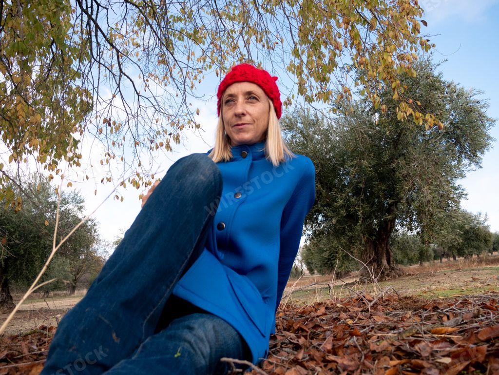 A shallow focus of a middle-aged blonde female wearing a blue coat and sitting on the ground in a park