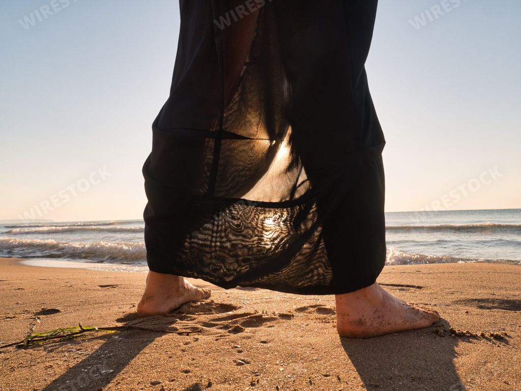 A female legs with brown dress standing on sandy beach touching sea water at sunset