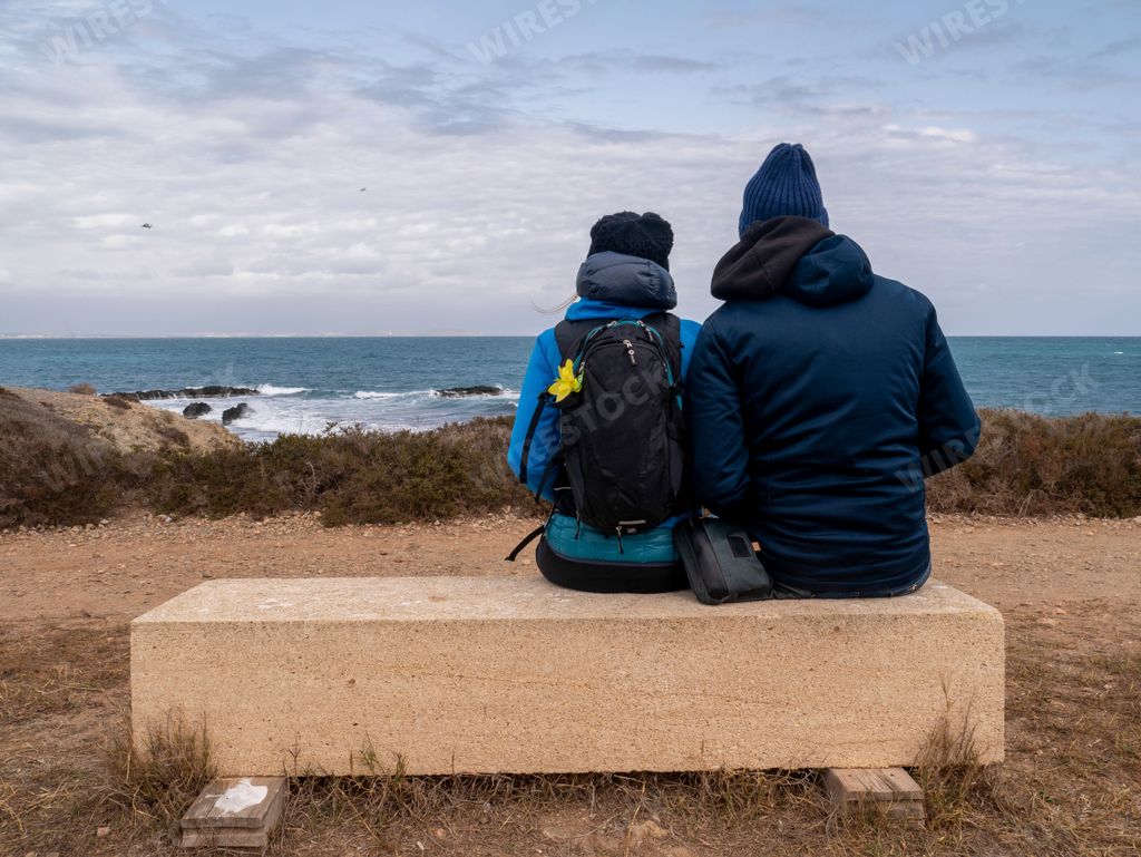 A back view of a couple sitting on a concrete bench in front of the sea on a cloudy day