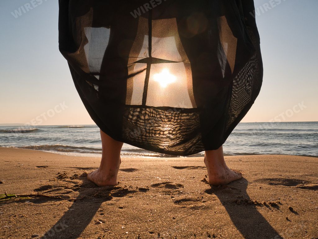 A female legs with transparent dress standing on sandy beach with sea water at sunset