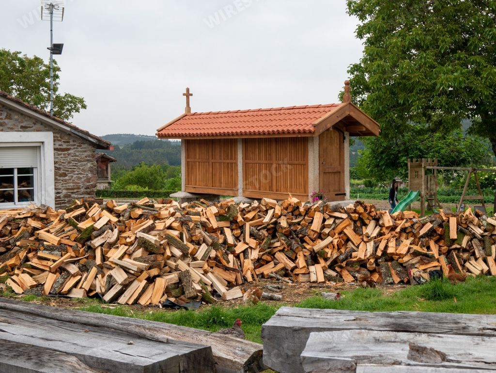 The piles of cut firewood on the grass and a narrow wooden cabin with a cross near.