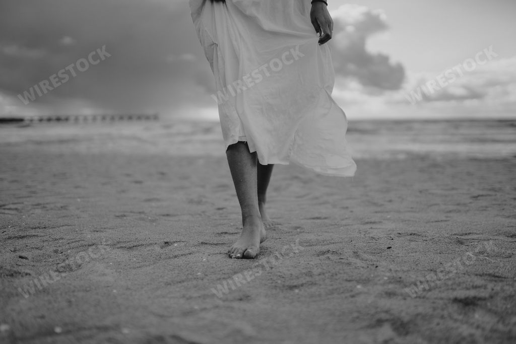 A greyscale shot of a beautiful lady's legs with a long white dress walking on the beach