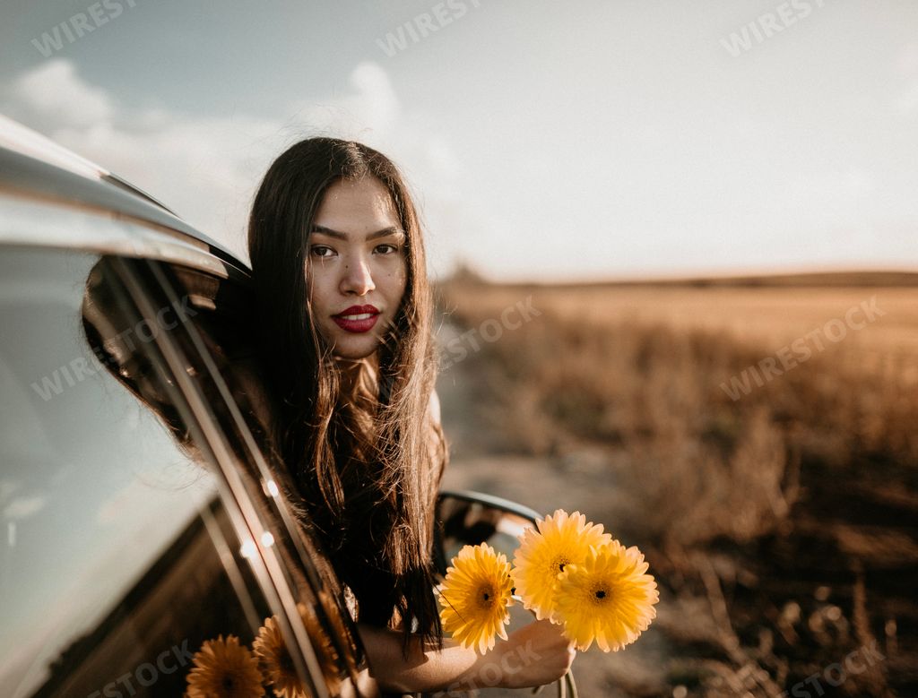 A cute brunette Hispanic woman posing flowers while hanging out the car window in the field