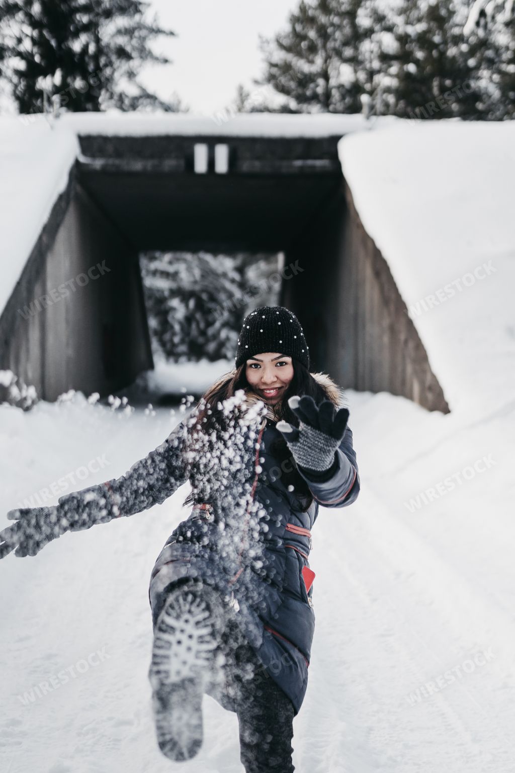 A young happy Hispanic woman wearing warm winter clothes playing with snow in a white snowy forest