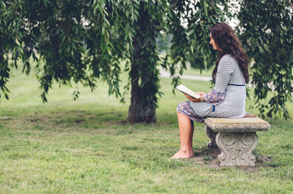 A Caucasian woman with long curly hair reading a bible at a park