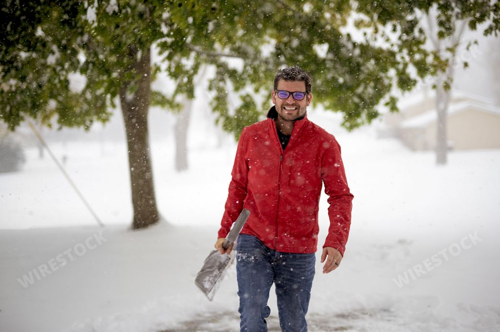 A male wearing a red jacket and walking in a snowy field while holding the snow shovel