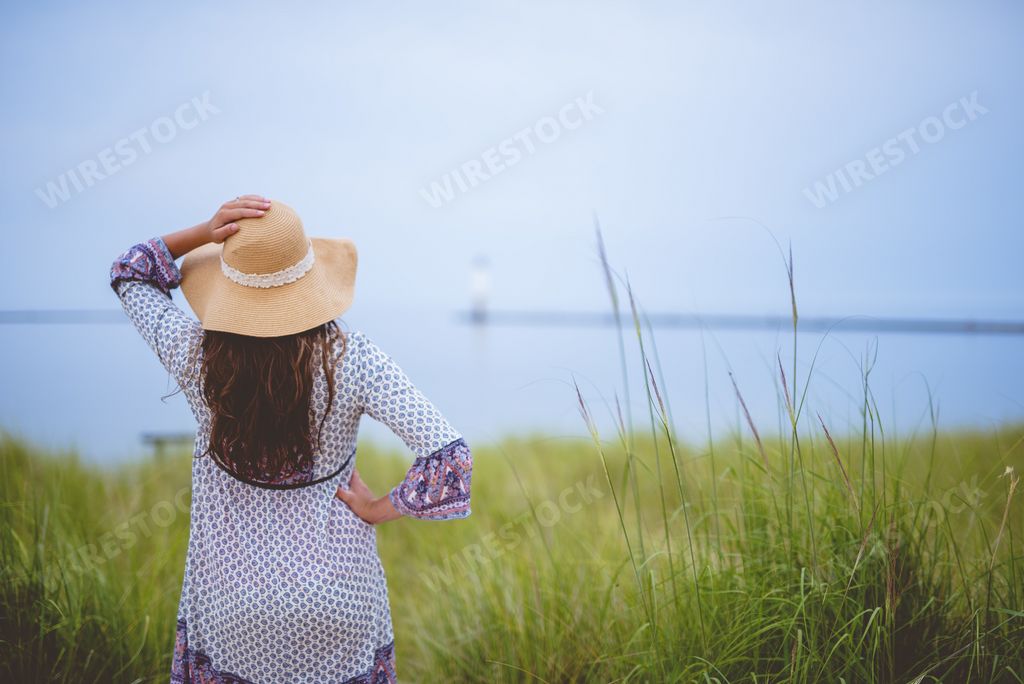 A female wearing a summer hat with her hand on the hip while standing in a grassy field shot from behind