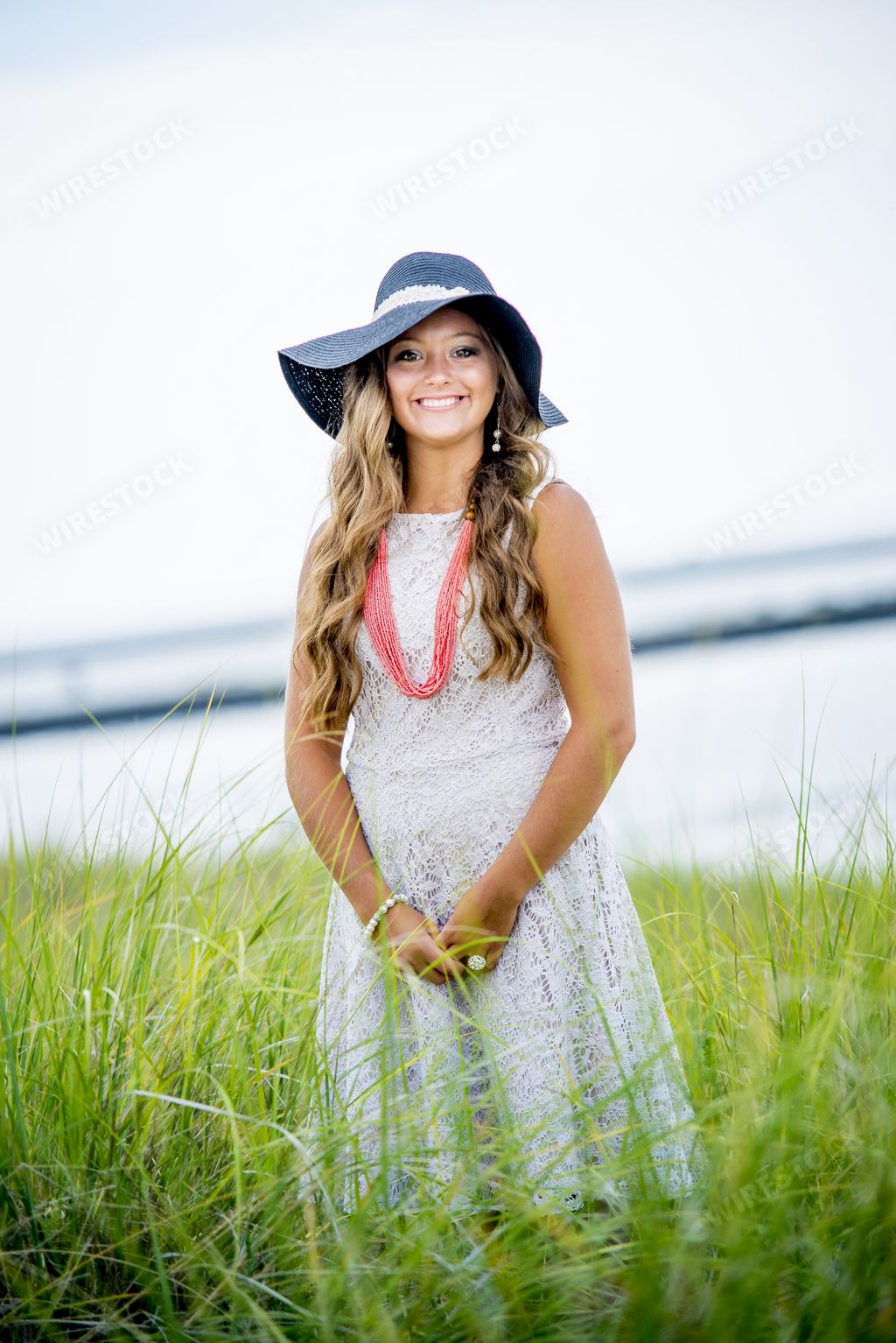 A blonde woman with a hat and a white dress standing in the garden with the beach on the background