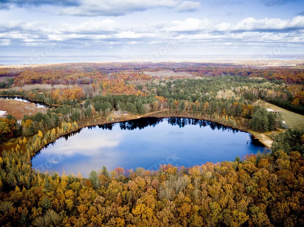 An aerial shot of a pond surrounded by different color trees under a blue cloudy sky