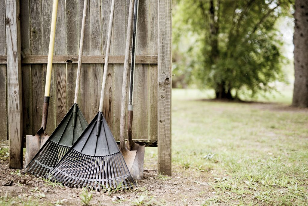 A closeup shot of two leaf rakes and shovels leaned against a wooden fence with a blurred background
