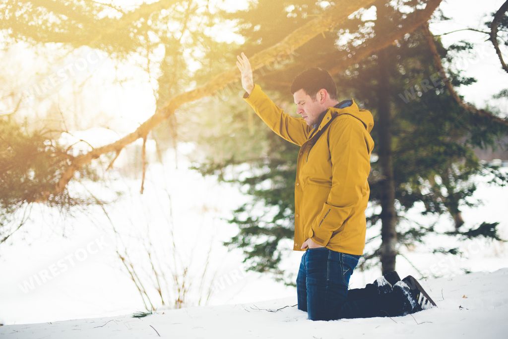 A male wearing a yellow snow jacket on his knees on the snow with his hand towards the sky - concept making amends