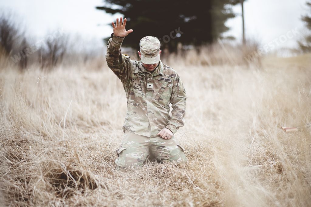 An American soldier kneeling in despair on a grass field with his hand raised above