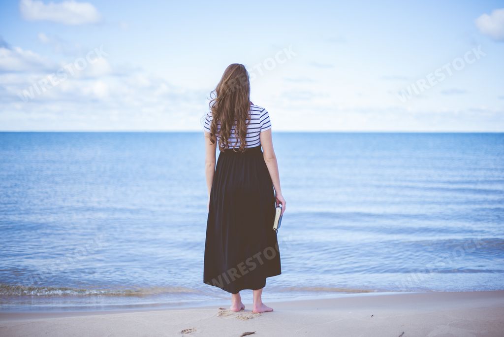 A female standing on a beach shore bare feet while holding the bible with a blurred background