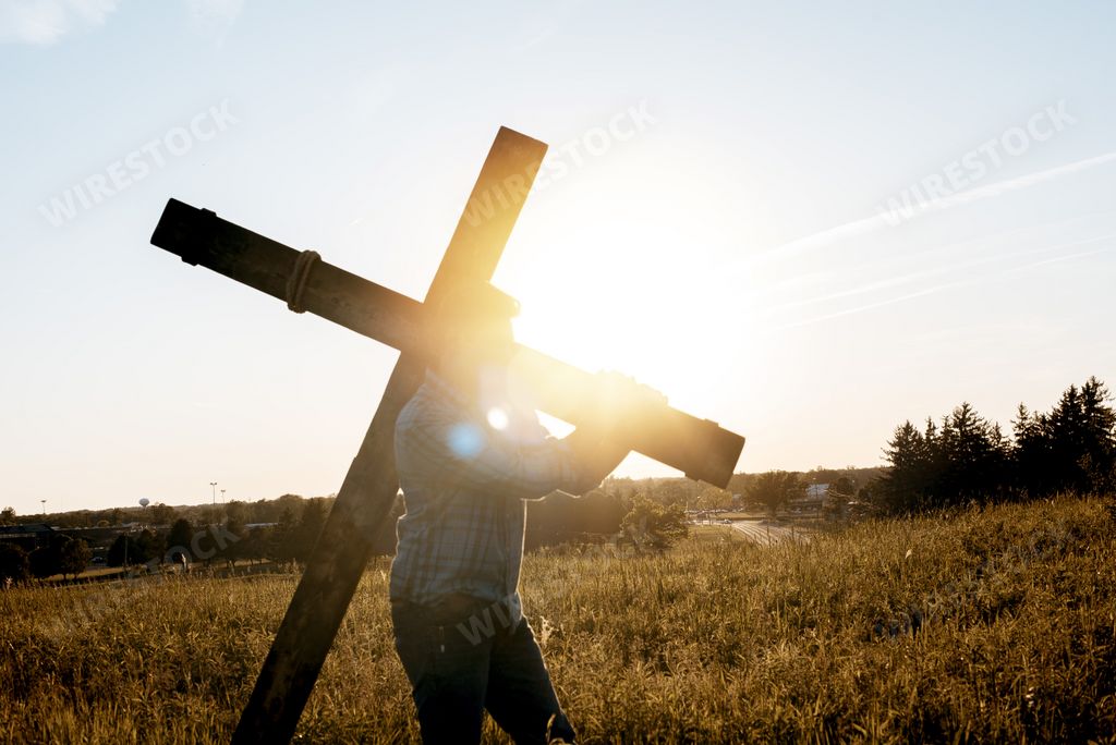 A closeup shot of a male carrying a wooden cross with the sun shining in the background