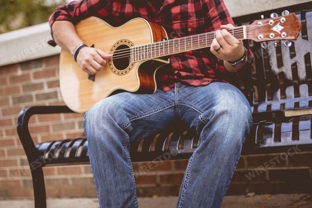 A male wearing a red and black flannel sitting on a bench playing the guitar