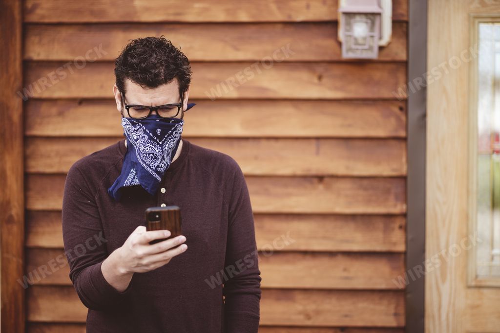 A male wearing glasses and a scarf as a face mask using his phone in front of a wooden wall