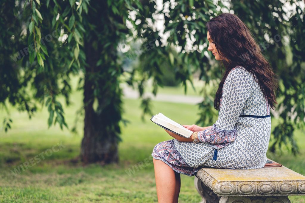 A Caucasian woman with long curly hair reading a bible at a park