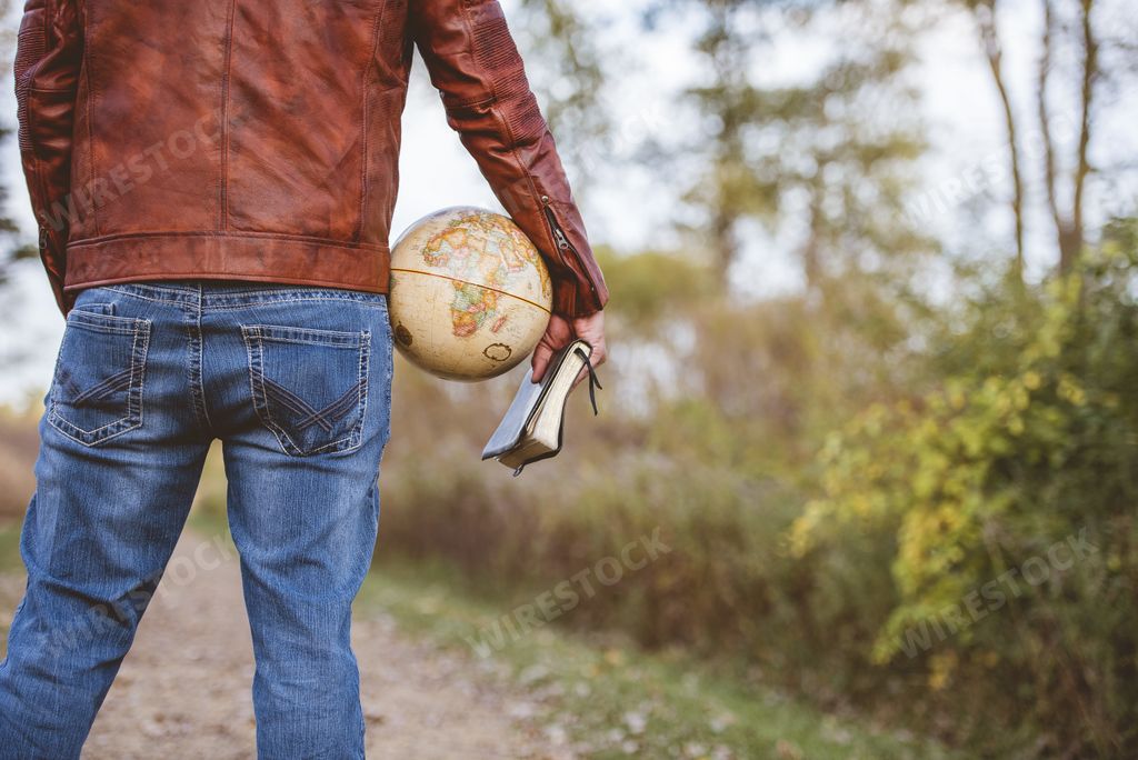 A male wearing a leather jacket and jeans holding a desk globe and the bible with a blurred background shot from behind