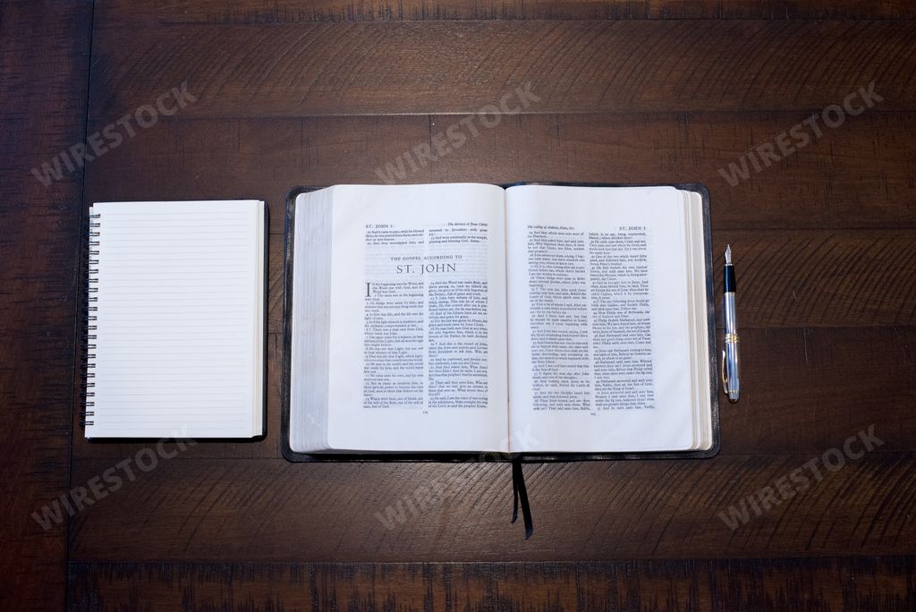 An overhead shot of an open bible in the middle of a notebook and a pen on a wooden surface