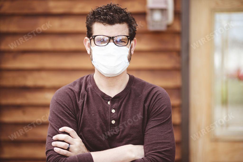 A male wearing glasses and a sanitary face mask with crossed arms in front of a wooden wall