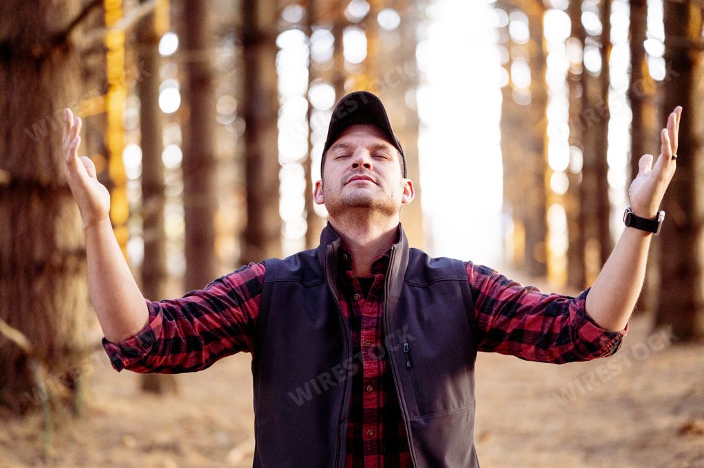 A Caucasian man praying in a forest with his hands raised towards the sky