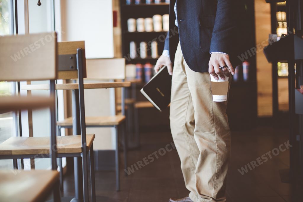 A closeup shot of a male holding his coffee and the bible while standing near a wooden table with a blurred background