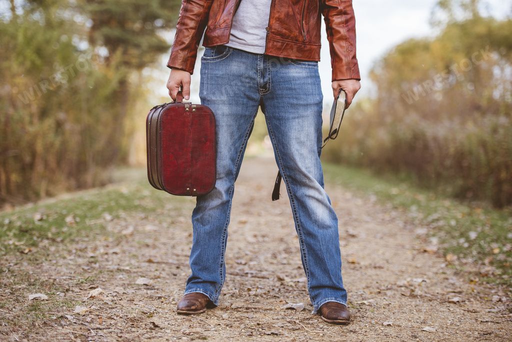 A male wearing a leather jacket and jeans standing on a pathway holding an old suitcase and the bible  - concept getting ready to travel