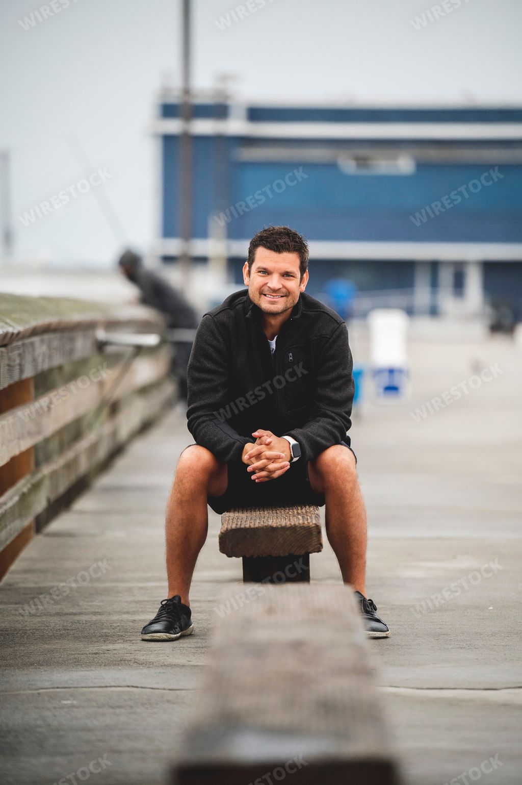 A vertical shot of a smiling man in a black jacket sitting on the bench and looking at the camera.