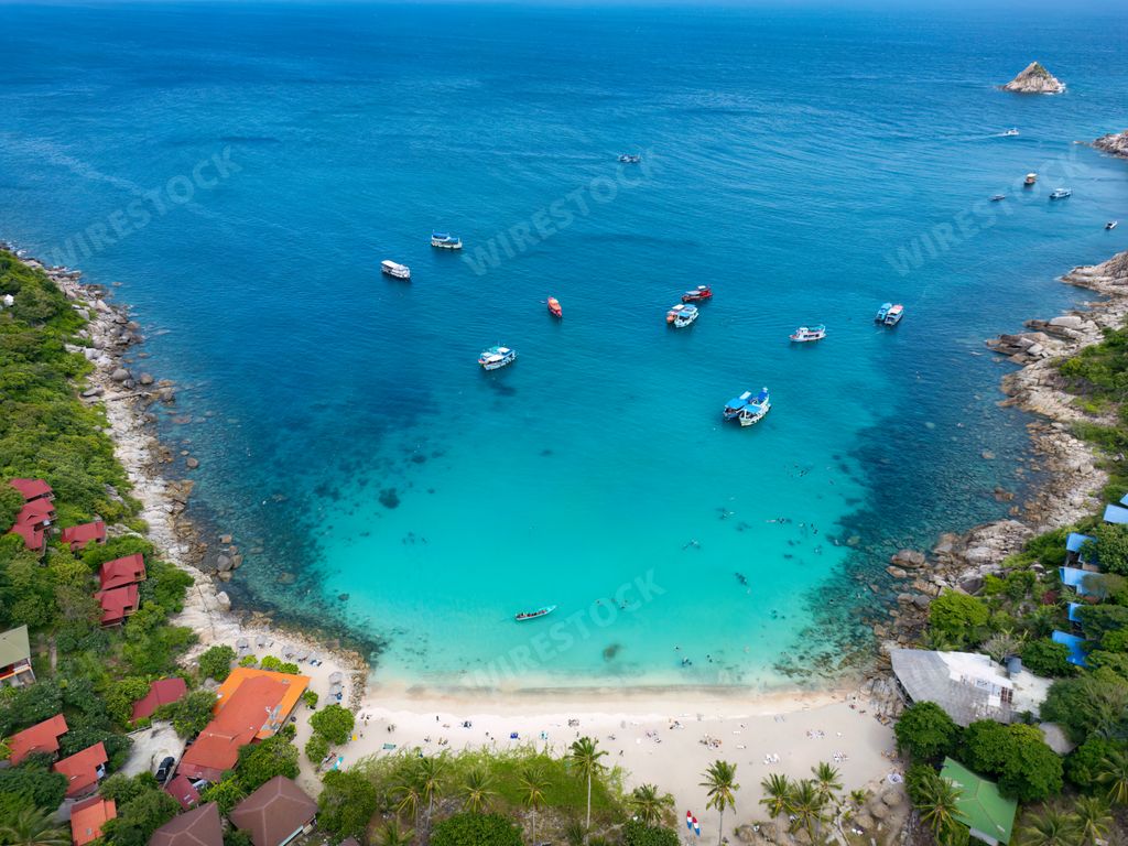 An aerial view of a beautiful tropical beach with clear blue water, boats, and lush greenery on Koh Tao Island, Thailand