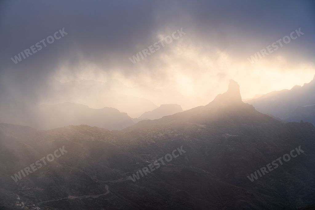 The Roque Bentayga peak in the Canary Islands covered with dark clouds with sunlight in the background