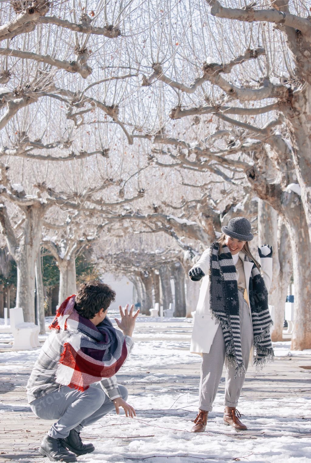 A vertical shot of a cute cheerful couple in a winter park: the lady throwing a snowball on the guy