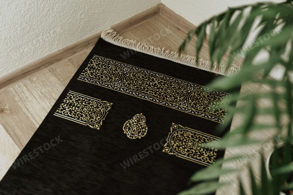 A top view of a black velvet prayer mat with a Kaaba pattern in the corner against a background of a house plant