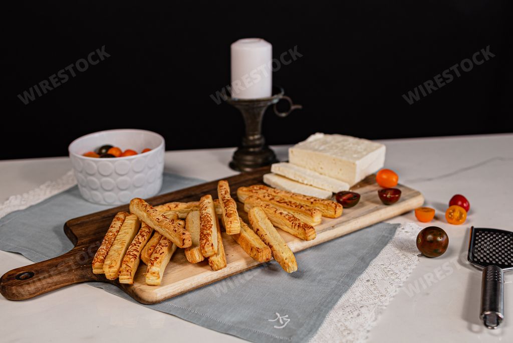 A pile of salted bread sticks on a wooden cutting board with some cheese and tomatoes