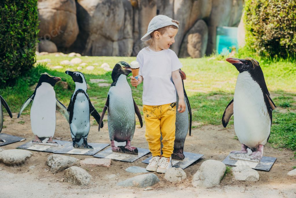 An adorable little boy holding icecream posing with little statues of penguins at a zoo