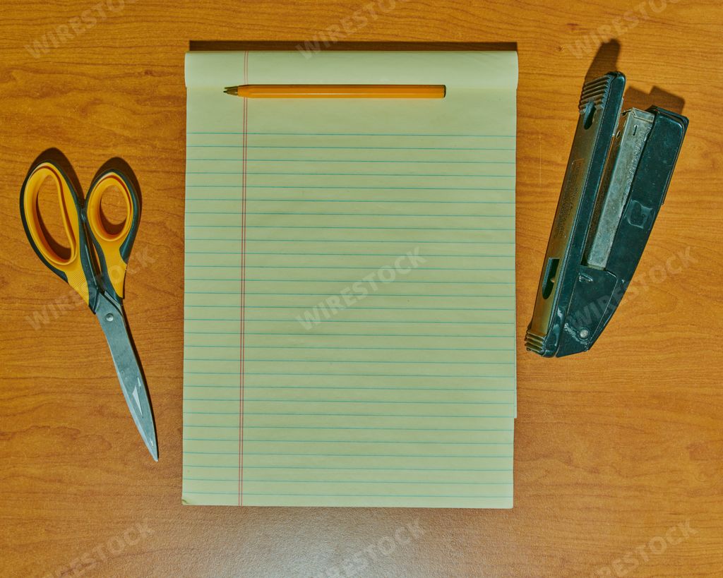 A top view of the blank lined open notebook, scissors, stapler, and pencil on the wooden desktop background