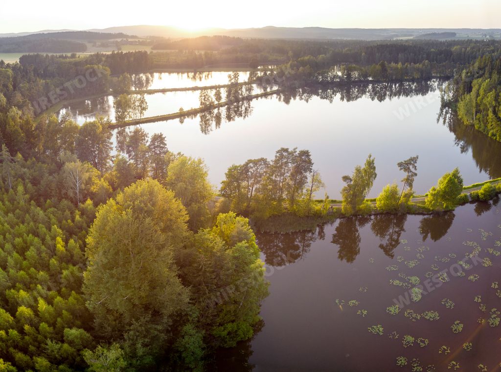 An aerial shot of a beautiful water pond surrounded by trees with hills and a sunset in the background