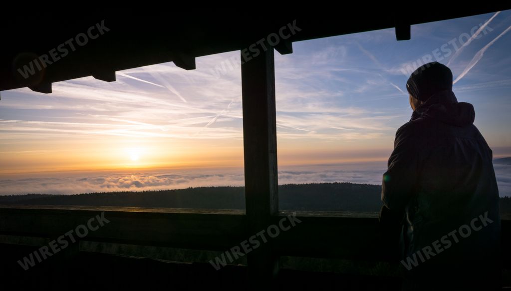 A silhouetted figure stands atop a mountain, gazing out into the horizon at sunset