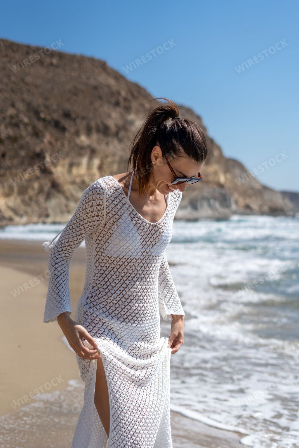 A Woman in white beach cover-up walking along a sandy beach with rocky cliffs and waves in the background.