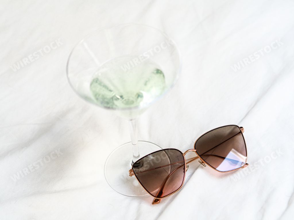 A top-view of glamorous sunglasses by a cocktail glass on white sheets